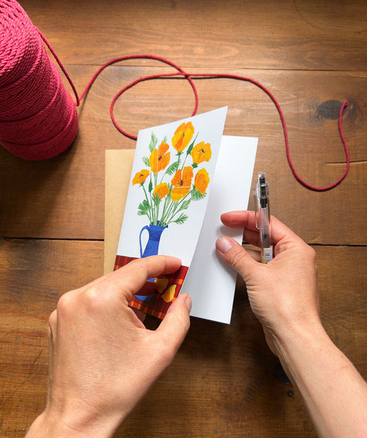 hands holding artist designed greeting card featuring bright poppies in a blue vase