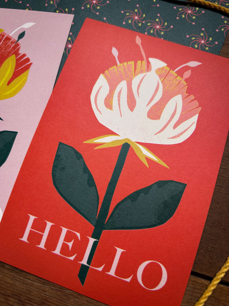 Red card with a white and pink flower illustration and 'HELLO' text on a wooden surface.
