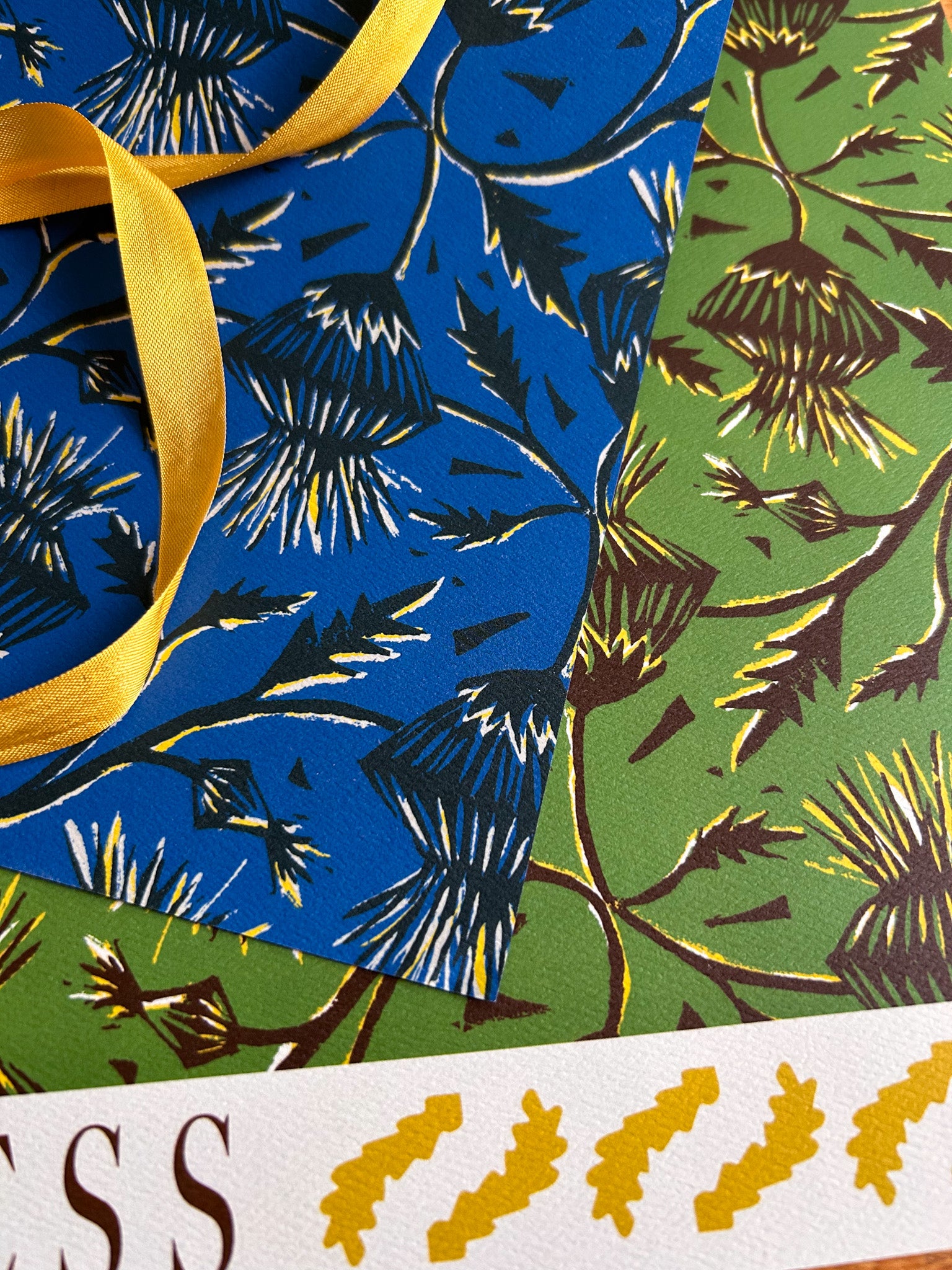 A double sided sheet of wrapping paper with a thistle flower pattern in green and blue colorways